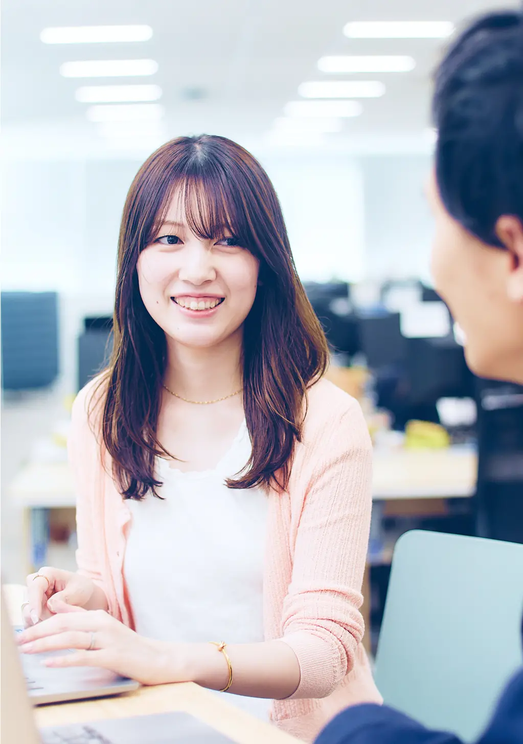 オフィスで同僚と会話をする、ピンクのカーディガンを着た女性社員。白いトップスと優しい笑顔が印象的なコミュニケーションシーン
