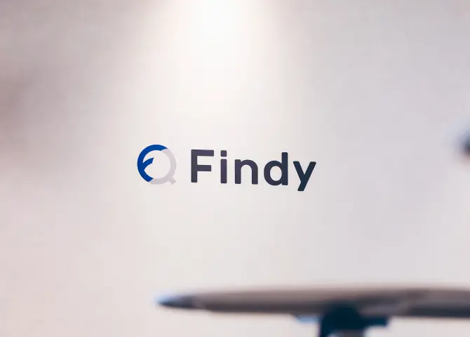 白い壁面に配置された、ブルーとグレーを基調としたFindyのロゴマーク。手前にぼかしで映るオフィス家具が奥行きを演出するコーポレートイメージ