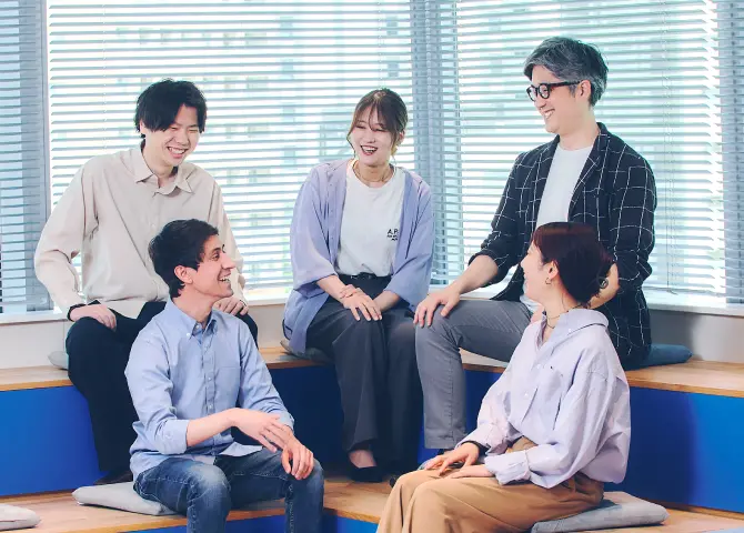 窓際のブルーのソファスペースで、カジュアルな服装の社員5名が和やかに会話を楽しんでいる様子