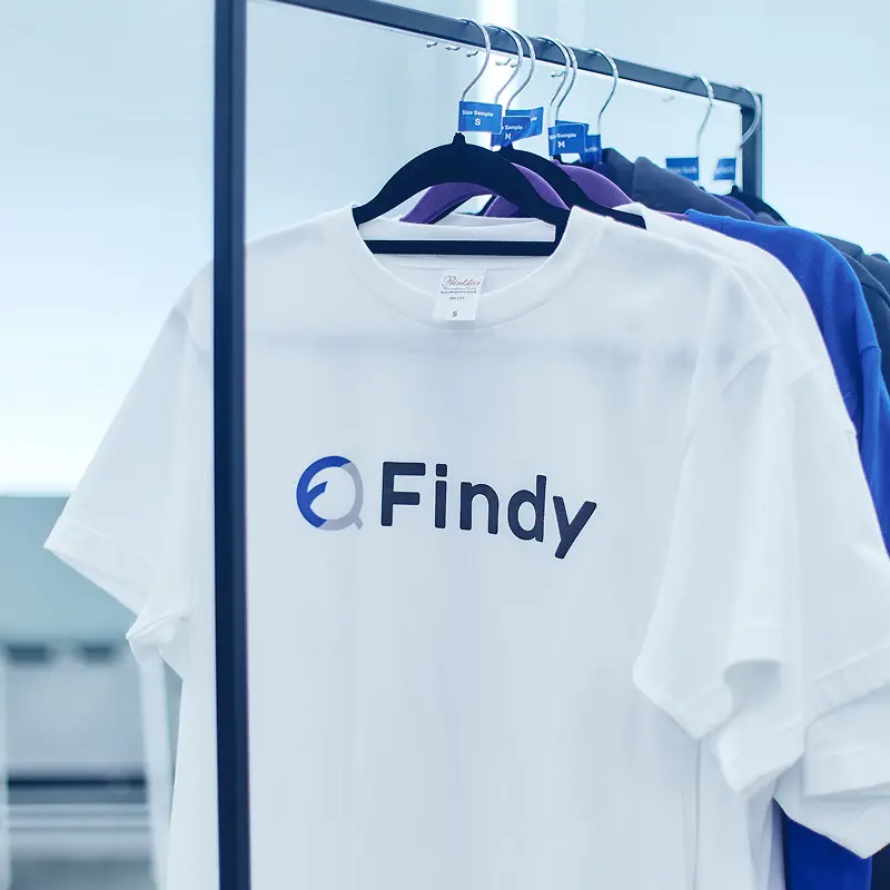 ハンガーラックに掛けられたFindyのコーポレートロゴ入り白とブルーのTシャツ。オフィシャルユニフォームとしての洗練されたデザイン