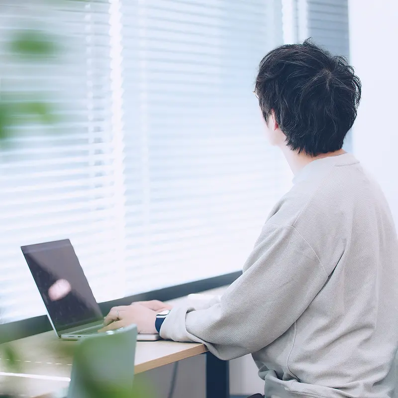 窓際のデスクでノートPCを操作する社員の後ろ姿。ベージュのニットを着用し、静かな環境で集中して作業に取り組む様子