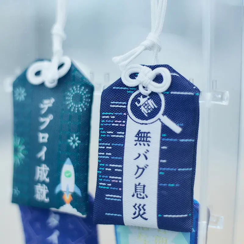 和風デザインの御守り風アイテム。「デプロイ成就」と「無バグ息災」の文字が刺繍された青と白の布製お守り