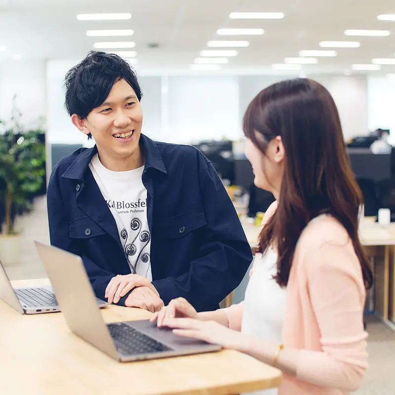 明るいオフィスで同僚と打ち合わせをする社員。ネイビーのジャケットとグラフィックTシャツを着用し、笑顔で会話を交わす様子