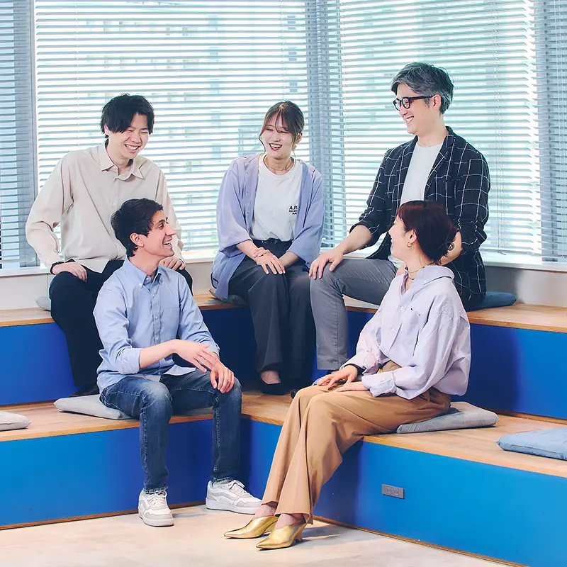 ブルーのステップ状ベンチに腰かけ、和やかに談笑する5名の社員。カジュアルな服装でリラックスした雰囲気の社内コミュニケーションシーン