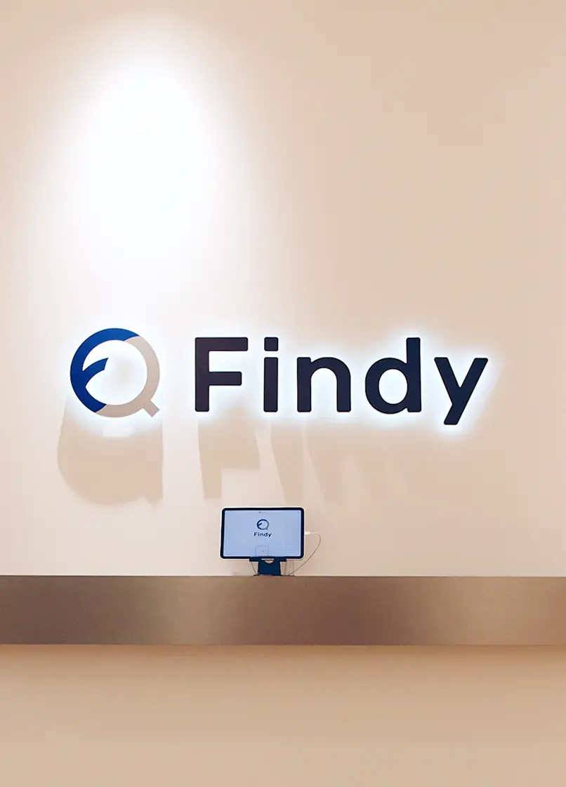 ベージュの壁面に設置された、バックライト付きのFindyロゴサイン。ブルーとグレーのコーポレートカラーが調和し、その下にはタブレット型の受付システムが配置された洗練されたエントリースペース