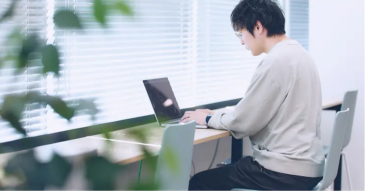 窓際のデスクでノートPCを操作する社員。ベージュのニットを着用し、観葉植物越しに撮影された集中して作業に取り組む横顔のショット
