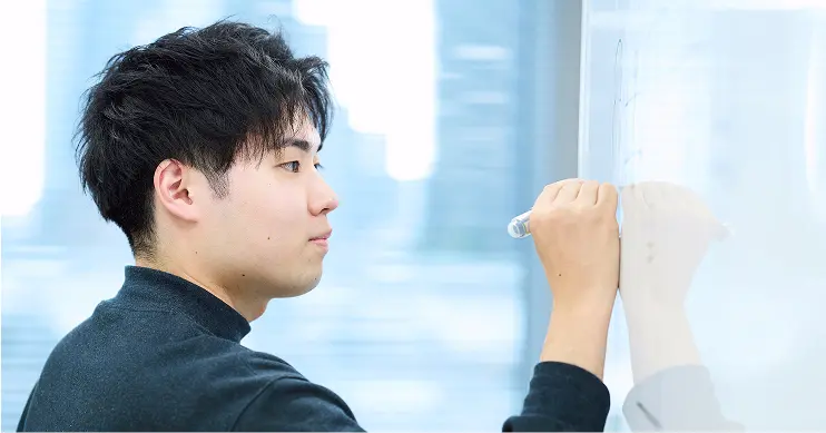 ホワイトボードに向かって企画を書き込む社員。黒のハイネックを着用し、都会的な景色を背景に、真剣な表情でアイデアを書き出す横顔のショット