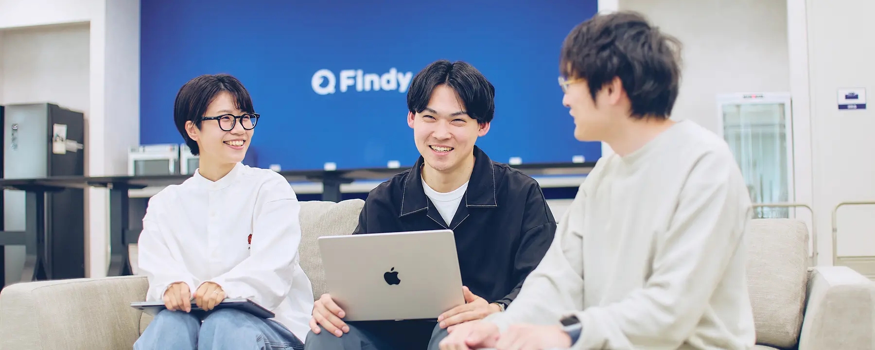 Findyのブルーのロゴウォールを背景に、ベージュのソファに座って和やかに会話を交わす3名の若手社員。白のスウェットやブラックのジャケットなどカジュアルな服装で、リラックスした雰囲気のミーティングシーン