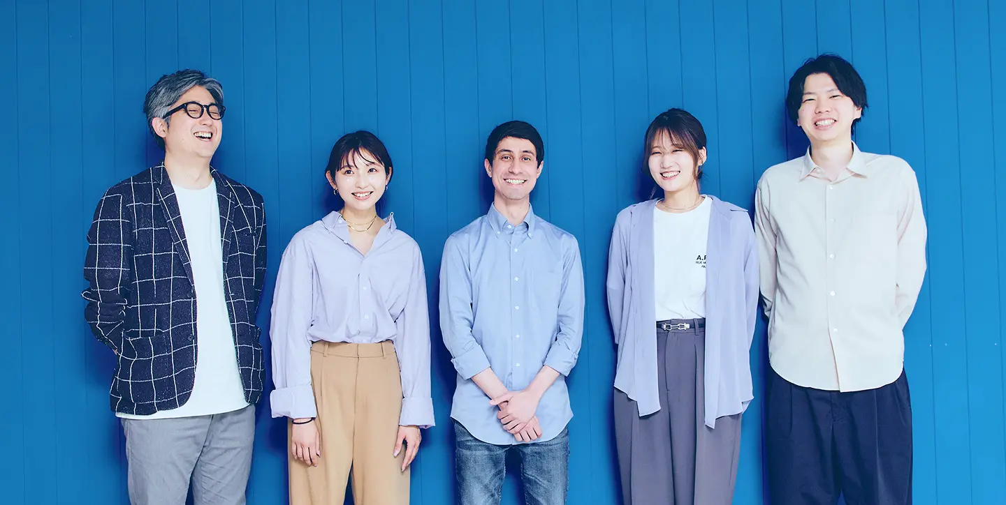 ブルーの壁を背景に並ぶ5名のFindyメンバー。左からチェック柄ジャケットの男性、ライラックシャツとベージュパンツの女性、ライトブルーシャツとデニムの男性、ライラックカーディガンとグレーパンツの女性、白シャツと黒パンツの男性が、それぞれ自然な笑顔で撮影に臨む集合写真