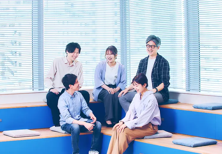 窓際のブルーのソファスペースで、カジュアルな服装の社員5名が和やかに会話を楽しんでいる様子