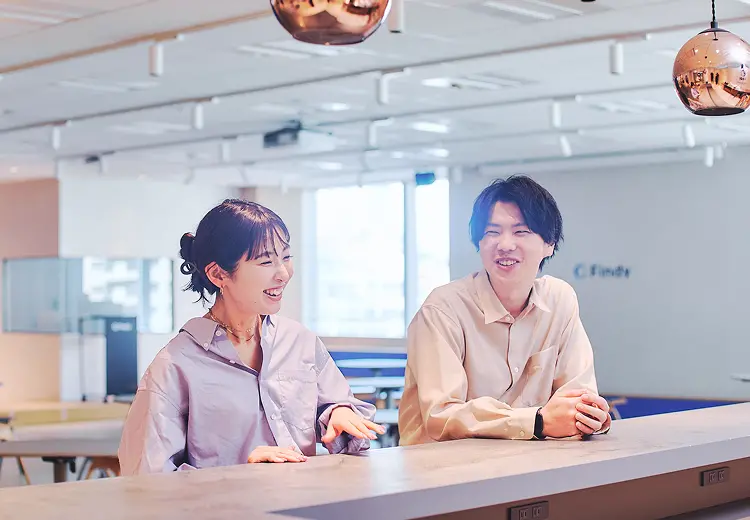 カウンターテーブルで和やかに会話を楽しむ2名の社員。温かみのある照明と明るい笑顔が印象的なオフィスシーン