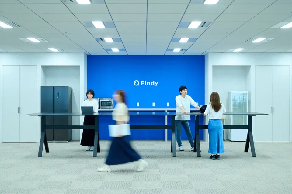 ブルーの壁面とFindyロゴを背景に、黒のハイテーブルで立ち仕事やコミュニケーションを取る社員たち。人の動きがモーションブラーで表現された開放的なオフィスシーン