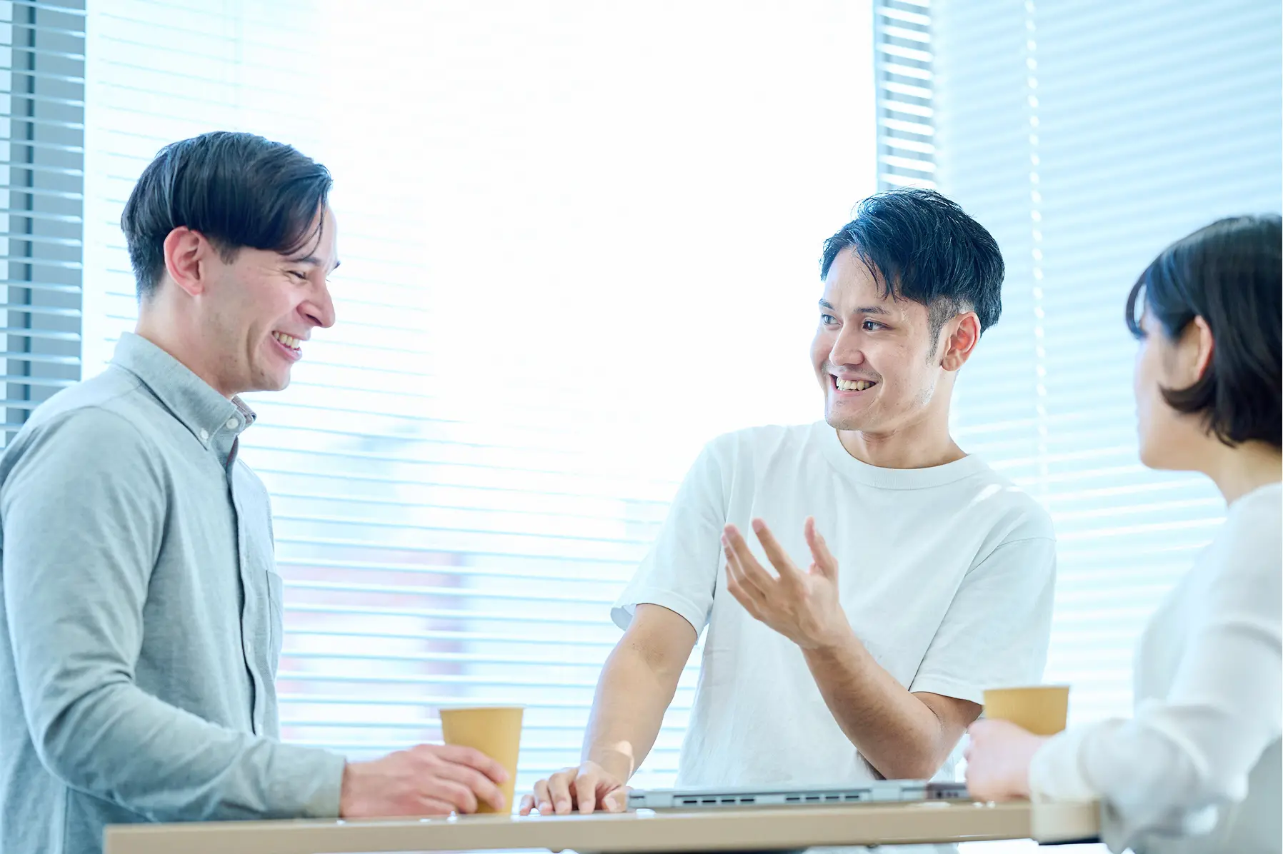 明るい窓際で、コーヒーを片手に活発な会話を交わす3名の社員。身振り手振りを交えた説明と笑顔から、フレンドリーな職場の雰囲気が伝わるシーン