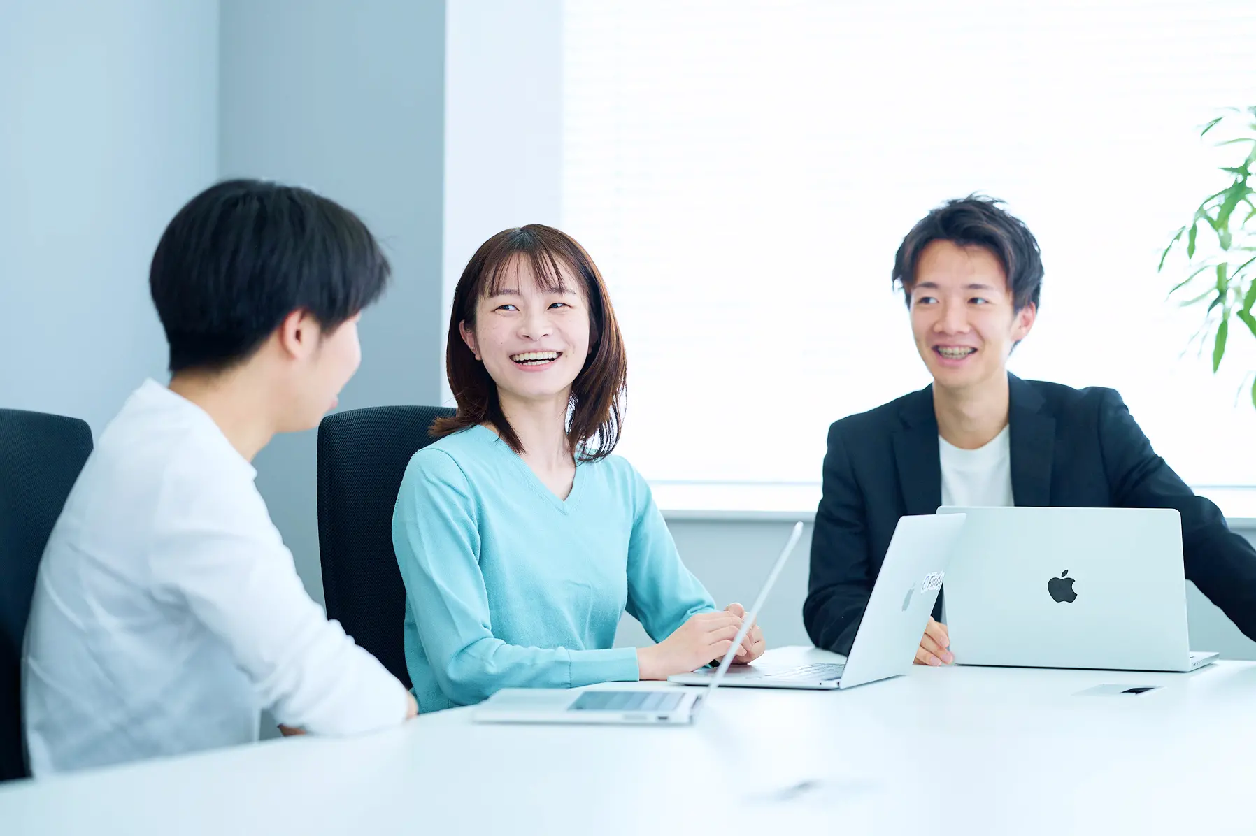 明るい会議室で、MacBookを前に和やかな雰囲気でミーティングを行う3名の社員。ミントグリーンのトップスを着た女性の笑顔が印象的なシーン