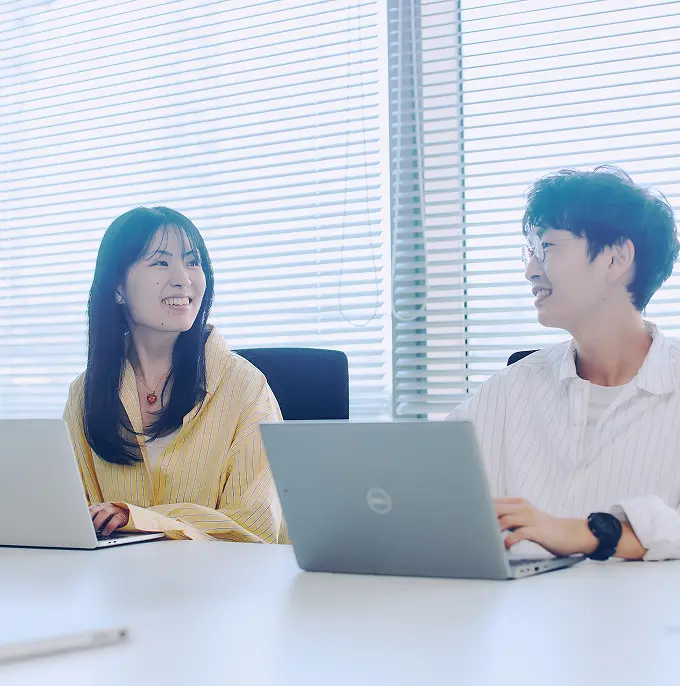 明るい窓際のデスクで、ノートPCを前に笑顔で会話を交わす2名の社員。イエローのストライプシャツを着た女性と白シャツの男性の自然な表情が印象的なシーン