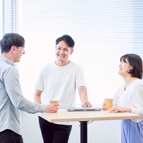 明るい窓際のハイテーブルで、コーヒーを片手に和やかに会話を楽しむ3名の社員。カジュアルな服装と自然な笑顔が印象的なオフィスシーン