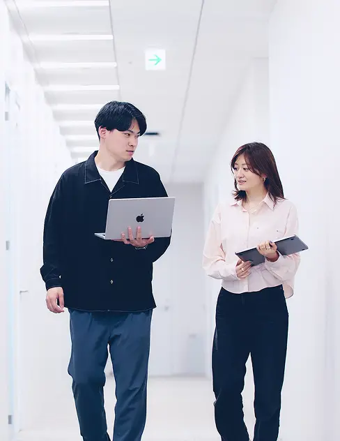 白を基調とした明るい廊下で、MacBookとタブレットを手に歩きながら打ち合わせをする2名の社員。ビジネスカジュアルな装いで真剣な表情を見せるシーン